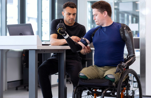 Man sitting at a desk helping a quadriplegic man in a wheelchair put on prosthetics  