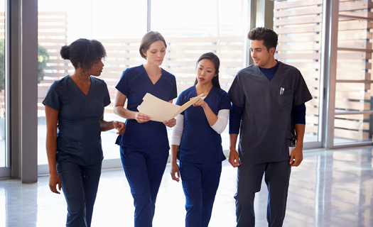 A group of diverse healthcare workers walking in a hallway with a large window behind them, discussing a file.