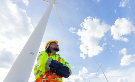 Utility worker standing beneath a wind turbine and smiling up toward a blue sky.